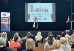 Elisabeth Gassner, Abteilungsleiterin des AMS Burgenland, steht auf einer Bühne und begrüßt die anwesenden Teilnehmer:innen